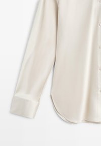 Camicia a maniche lunghe color crema realizzata in tessuto morbido, con orlo curvo, chiusura con bottoni e dettagli delle maniche arrotolate.