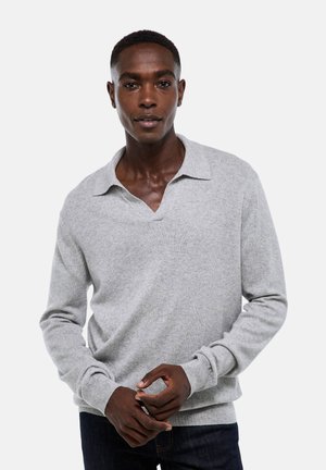 Homme portant un pull en maille gris clair à manches longues avec col, se tenant contre un fond blanc uni, les mains légèrement jointes.