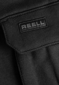 Black fabric close-up featuring a REELL brand label with raised lettering. The texture is smooth with visible stitching details.