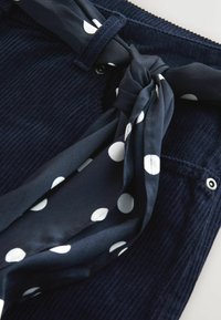 Granatowa tkanina sztruksowa o fakturowanym wykończeniu, z białym krawatem szalika w kropki zawiązanym w supeł przy pasku. Metalowy guzik jako detal.