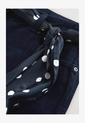 Granatowa tkanina sztruksowa o fakturowanym wykończeniu, z białym krawatem szalika w kropki zawiązanym w supeł przy pasku. Metalowy guzik jako detal.
