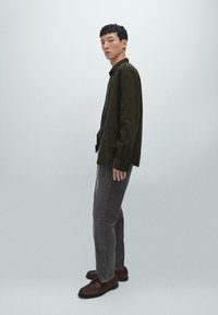 Camicetta bottonata verde scuro sopra una t-shirt blu scuro, jeans grigi a gamba larga e scarpe marroni, posata davanti a uno sfondo grigio chiaro.