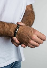 Žalias pintas paracordo apyrankė su metaliniu sidabro užsegimu. Nešiojama ant riešo, matoma plaukų tekstūra. Paprastas ir patvarus dizainas.