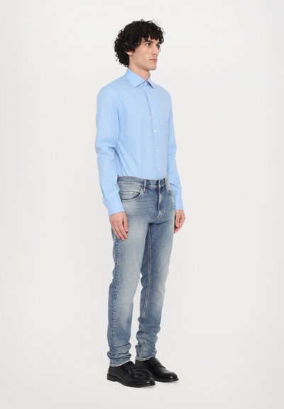 Lichtblauwe overhemd met lange mouwen en knopen, gecombineerd met vervaagde blauwe jeans. Model draagt zwarte instappers. Eenvoudig, passend ontwerp.