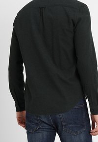 Dunkelgrünes Langarmhemd mit geknöpftem Kragen, das einen plissierten Rücken und einen abgerundeten Saum aufweist, kombiniert mit blauen Jeans.