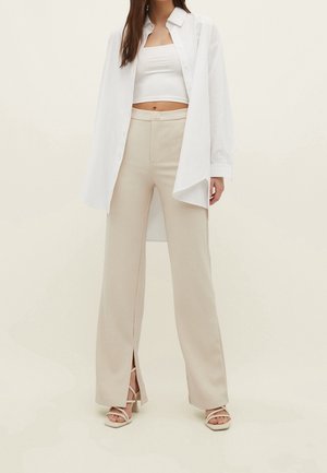 Femme portant un pantalon beige à jambes larges avec fentes devant, un top court blanc, une chemise blanche oversize boutonnée et des sandales blanches à talons et brides.