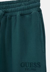 Mörk teal joggers i mjukt tyg, med elastisk midja, sidfickor och broderad "GUESS EST. 1981"-logga.