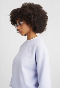 Lila pulóver négyzet alakú folttal a mellkason, kerek nyakkivágással és bordás mandzsettákkal. A modellen árnyékolt szemüveg van, és göndör haja van.