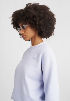 Lilafarget genser med en firkantet lapp på brystet, med rund hals og ribbestrikkede mansjetter. Modellen har på seg fargede briller og har krøllete hår.