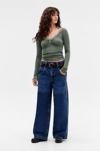 Long-sleeve olive green ribbed top with lace trim, paired with wide-leg blue denim jeans and a black belt featuring gold studs.