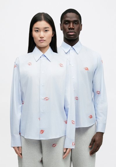 Light blue button-up shirts feature red lipstick kiss prints, with long sleeves and a classic collar. The fabric appears smooth with a slight sheen.
