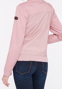 Veste rose pâle à texture lisse, avec une écusson marron sur l'épaule gauche et une taille cintrée, associée à un jean bleu foncé.