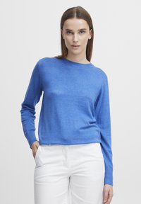 Pull en tricot bleu à manches longues et à col rond, affichant une texture lisse et un design minimaliste. Associé à un pantalon blanc.