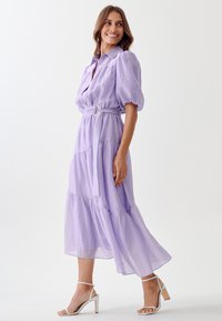 Lavender, tiered shirt dress with puff sleeves, belted waist, and a flowing hem. Accompanied by strappy white high heels for a complete look.