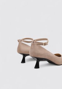 Talons en suède beige avec un talon noir courbé et une lanière de cheville. Le design élégant présente un bout arrondi et une texture lisse.