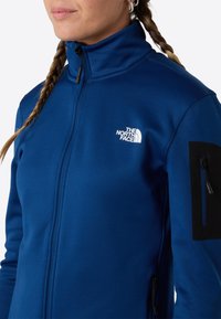Modrá fleecová bunda s vysokým límcem, černými akcenty na rukávě a kapsou na zip. Na bundě je bílé logo "The North Face".