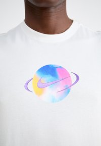 Balts kokvilnas T-krekls ar apļveida, daudzkrāsainu grafiku, kurā ir purpursarkans Nike zvīņas simbols un apkārtējie apļi purpursarkanā un maigu pastelīgu toņu krāsās.