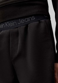 Pantaloni sportivi neri con texture morbida, tasche frontali, una vita elasticizzata a coste con logo "Calvin Klein Jeans" e un dettaglio di branding discreto.
