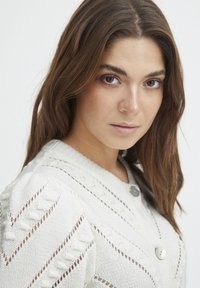 Cardigan en tricot de couleur beige clair avec des motifs ajourés, présentant un col rond et deux grands boutons à l'avant. Texture douce et design ajusté.