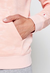 Sudadera con capucha rosa claro hecha de tela suave, con un bolsillo tipo canguro y puños acanalados. El logo en la manga añade un detalle.