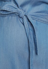Svetle modrá džínovina se zavázanou mašlí v pase. Textura vypadá hladce s mírným leskem a obsahuje minimalistické designové prvky.