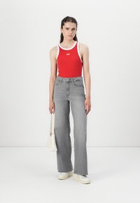 Rotes ärmelloses Oberteil mit weißem Saum und "GAP"-Logo, kombiniert mit hoch taillierten grauen Schlagjeans und weißen Sneakers. Hält eine weiße Handtasche.