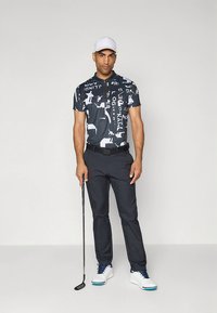 Mørk mønstret polo skjorte med hvite grafiske elementer, kombinert med mørke bukser. Tilbehør inkluderer hvit caps og lyse sko, holder en golfkølle.