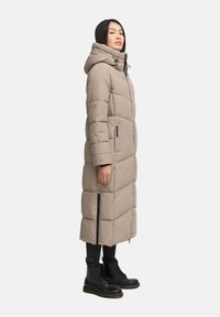 Abrigo acolchado largo beige con capucha, diseño de acolchado, bolsillos con cremallera y aberturas laterales, combinado con botas negras de cordones.