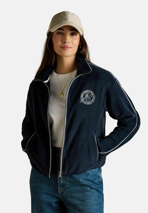 Woman wearing beige cap, navy zip-up jacket with white piping and logo, white shirt, gold pendant necklace, and blue jeans, hands in pockets.