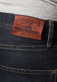 Etiqueta de cuero marrón en unos vaqueros de mezclilla azul oscuro con "Jack & Jones Vintage Denim" y un logotipo de pantera bordado en la cintura, encima del bolsillo trasero.