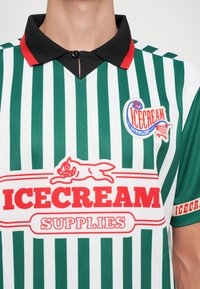 Groene en witte gestreepte polo met een zwarte kraag, voorzien van een rood logo en een afbeelding van een kat. Korte mouwen met een accentband.