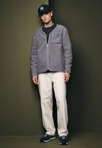 Giacca in pile grigia con zip e tasca, t-shirt nera, pantaloni beige chiaro e sneakers blu scuro, posizionati su uno sfondo verde tenue.