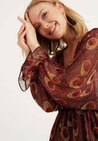 Langärmliges, durchsichtiges Kleid mit einem roten und goldenen Kreis-Muster auf dunklem Hintergrund. Elastischer Bund; ausgestellte Manschetten. Schwarze, runde Ohrringe.