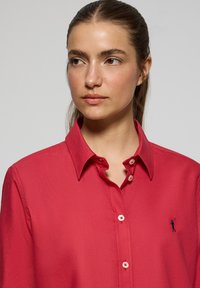 Camicia rossa con bottoni, colletto classico, con logo ricamato in blu navy. Tessuto liscio e vestibilità classica con bottoni a contrasto.