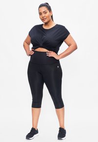 Zwarte sporttop met een geknoopte voorkant, gecombineerd met hoge zwarte capri-leggings. Model draagt zwarte sportschoenen.
