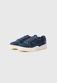 Blå lærredssneakers med gule gummisåler, hvid mellemsål og matchende snørebånd. Har kontrasterende syninger og teksturerede detaljer.