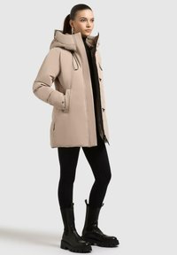 Cappotto puffer beige con cappuccio, dotato di zip frontale, tasche laterali e dettagli neri. Abbinato a leggings neri e stivali neri chunky.