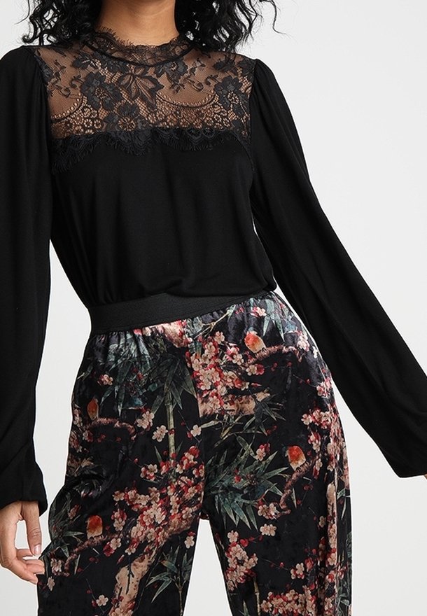 Czarna bluzka z długimi rękawami posiada przezroczysty koronkowy karczek ozdobiony kwiatowymi wzorami, zestawiona z luźnymi czarnymi spodniami w kwiaty.