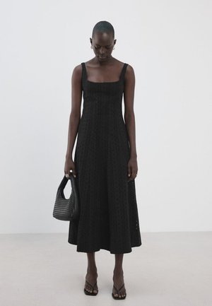 Kobieta w czarnej, bezrękawnikowej sukience midi z wzorem ażurowym, trzymająca czarną plecioną torebkę i nosząca czarne klapki.