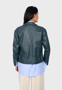 Tmavě modrá kožená bunda s límcem a dlouhými rukávy, s hladkou texturou a těsným střihem, nošená přes světle modrou košili.