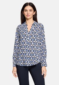 Blouse à manches longues blanche avec un motif ondulé bleu et noir, dotée d'un col et d'un devant à boutons, confectionnée en tissu léger.