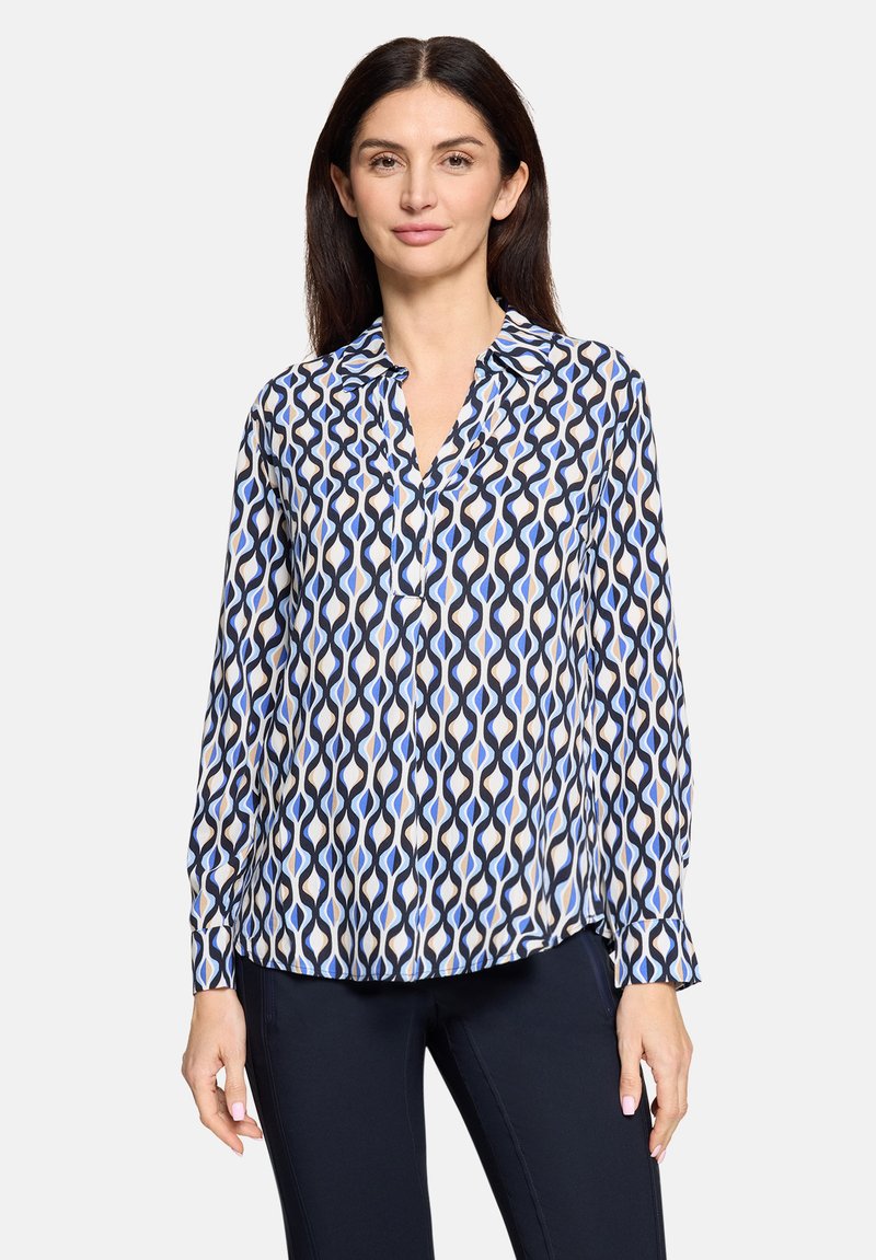 Blouse à manches longues blanche avec un motif ondulé bleu et noir, dotée d'un col et d'un devant à boutons, confectionnée en tissu léger.