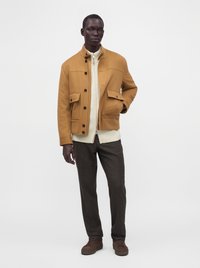 Homme portant une veste beige par-dessus un pull zippé crème, un pantalon foncé et des chaussures marron, debout devant un fond blanc uni.
