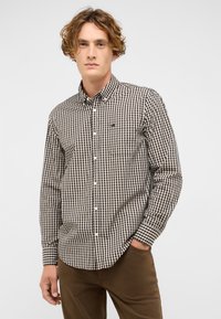 Junger Mann mit lockigem Haar, der ein knöpfbares schwarz-beiges kariertes Hemd und braune Hose trägt, eine Hand in der Tasche, neutraler Hintergrund.