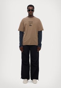 T-shirt marrone a maniche corte con testo frontale, sovrapposto a una maglietta a maniche lunghe blu scuro. Pantaloni neri a gamba larga, occhiali da sole e scarpe beige.