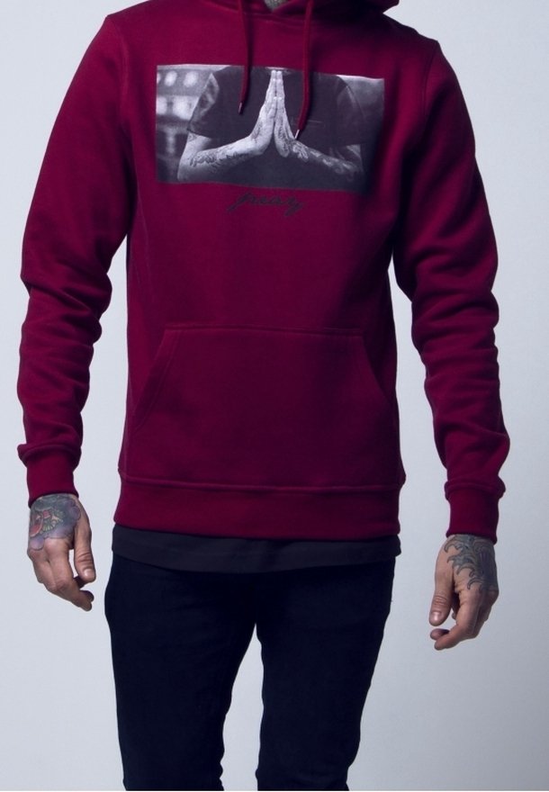 Personne portant un sweat à capuche bordeaux avec une image en noir et blanc de mains tatouées en position de prière sur la poitrine, mains visibles, debout.