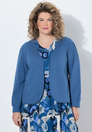 Frau mit lockigem Haar, die ein blaues Blumenkleid und eine einfarbige blaue Jacke trägt, vor einem hellen Hintergrund stehend.