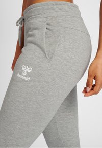 Pantalones de chándal grises hechos de tela suave, con una cinturilla elástica ajustable, bolsillos laterales y un logo blanco en el muslo. Textura suave.