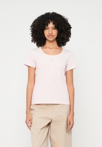 Kortsirviga kergete roosade varrukatega pluusidega naine lokkis juustega, seismas ühevärvilise valge tausta ees, seljas beežid püksid.