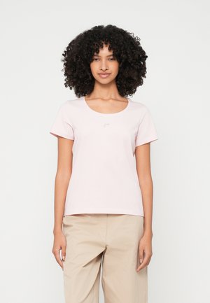 Göndör hajú nő világos rózsaszín, rövid ujjú pólót és bézs nadrágot visel, egy sima fehér háttér előtt áll.